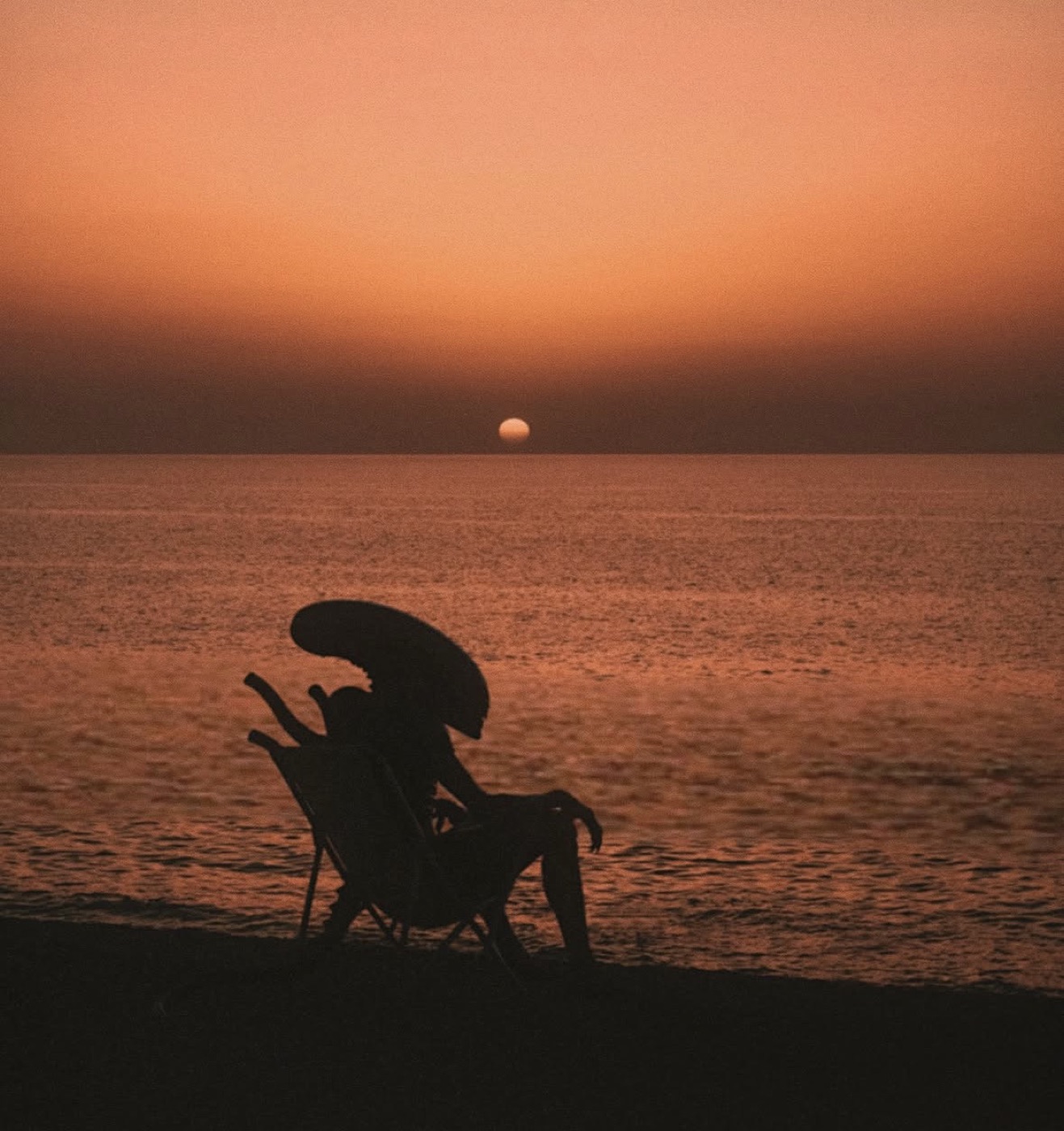 Silhouette of a xenomorph relaxing in a beach chair at sunset.